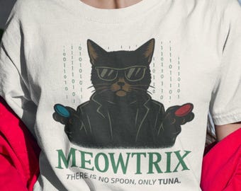 May include: Off-white t-shirt featuring a black cat wearing sunglasses and a black suit, with the text "MEOWTRIX" and "THERE IS NO SPOON, ONLY TUNA." The cat is holding a red and blue pill. The shirt is worn by a person.
