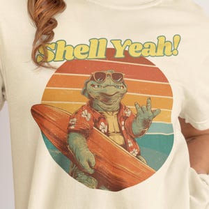 Könnte beinhalten: Cremefarbenes T-Shirt mit einem Retro-Grafikdesign einer Schildkröte mit Sonnenbrille und Hawaiihemd, die ein Surfbrett hält. Der Text "Shell Yeah!" ist in einem Vintage-Schriftzug über der Schildkröte angeordnet. Das Design ist vor einem Sonnenuntergangshintergrund.