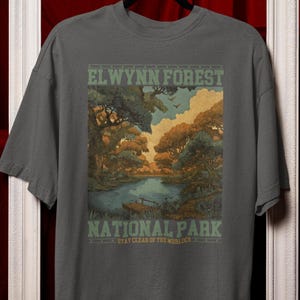 Könnte beinhalten: Graues T-Shirt mit einer malerischen Illustration des El Wynn Forest Nationalparks. Das Design umfasst einen See, Bäume und den Text "EL WYNN FOREST NATIONAL PARK" in Grün und Weiß. Das Shirt hat einen Rundhalsausschnitt und kurze Ärmel.
