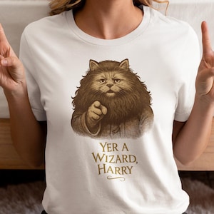 Puede incluir: Camiseta blanca con una ilustración en tonos sepia de un gato señalando. El texto "Yer a Wizard, Harry" está debajo de la imagen en un tono sepia a juego. La camiseta está hecha de un material suave.