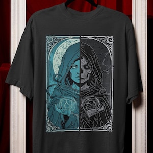 May include: Dark gray t-shirt featuring a split design. One side depicts a hooded figure in blue, holding a glowing orb. The other side shows a skeletal figure in black, also holding an orb. The design is framed with ornate details.