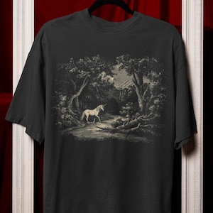 May include: Black t-shirt with a sepia-toned illustration of a unicorn in a forest. The design shows a unicorn walking on a path surrounded by trees, creating a mystical and enchanting scene.