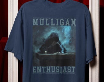May include: Navy blue t-shirt featuring the text "MULLIGAN ENTHUSIAST" in a gray, block font. The shirt has a graphic of a hooded figure in a dark, smoky setting. The t-shirt is hanging on a black hanger.
