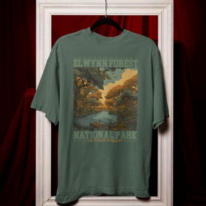May include: Green t-shirt featuring a scenic illustration of a forest and lake with the text "ELWYNN FOREST NATIONAL PARK". The design includes trees, water, and a sky with clouds. The shirt is hanging on a black hanger.