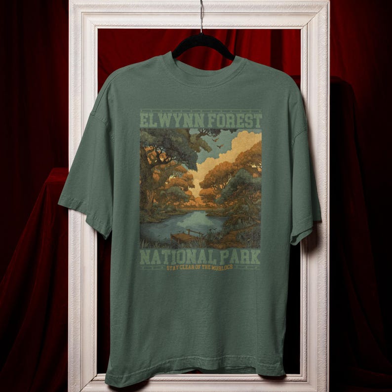 May include: Green t-shirt featuring a scenic illustration of a forest and lake with the text "ELWYNN FOREST NATIONAL PARK". The design includes trees, water, and a sky with clouds. The shirt is hanging on a black hanger.