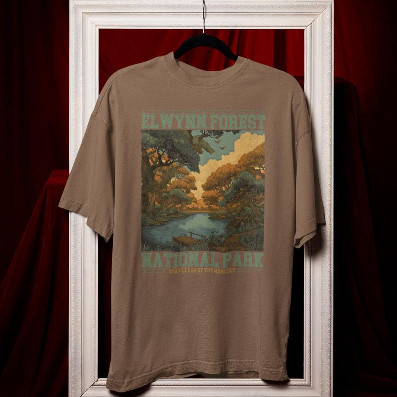 May include: A brown t-shirt featuring a scenic illustration of a forest and lake. The text "EL WYNN FOREST NATIONAL PARK" is printed in green. The shirt is displayed on a black hanger against a red backdrop.