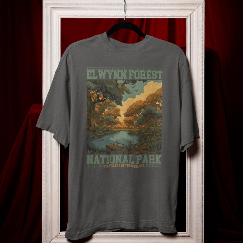 May include: Gray t-shirt featuring a scenic illustration of El Wynn Forest National Park. The design includes a lake, trees, and the text "EL WYNN FOREST NATIONAL PARK" in green and white. The shirt is a short-sleeved crew neck.