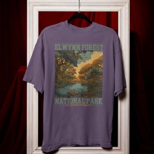 May include: A purple t-shirt featuring a scenic illustration of a forest and lake. The text "ELWYNN FOREST NATIONAL PARK" is printed in green and white. The shirt has short sleeves and a crew neck.