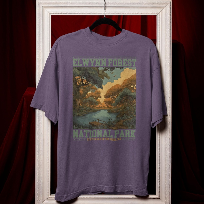 May include: A purple t-shirt featuring a scenic illustration of a forest and lake. The text "ELWYNN FOREST NATIONAL PARK" is printed in green and white. The shirt has short sleeves and a crew neck.