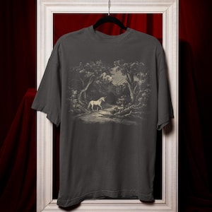 May include: Dark grey t-shirt with a black and white illustration of a unicorn in a forest scene. The image shows trees, a stream, and a building. The t-shirt is displayed on a black hanger against a red backdrop.