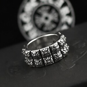 May include: A silver-toned metal ring featuring two stacked rows of square segments, each embossed with a detailed cross emblem. The band has a polished finish and a consistent width, showcasing a repeating pattern of crosses around the entire circumference.