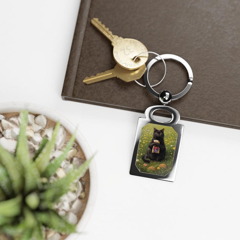 May include: A silver-toned rectangular keychain featuring a central image of a black cat sitting in a field of yellow dandelions and holding a small bag of snacks. The keychain includes a circular metal ring attached to two brass-colored keys.