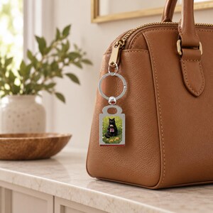 May include: A silver-toned metal keychain featuring a rectangular pendant with a printed image of a black cat sitting in a field of yellow flowers. The keychain is attached to the gold-toned zipper pull of a brown leather handbag.
