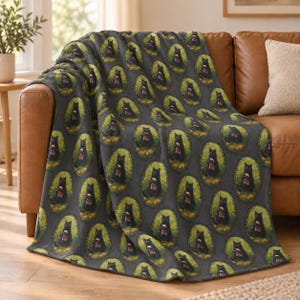 May include: A dark gray fleece throw blanket featuring a repeating pattern of black cats sitting inside oval frames. Each cat is depicted holding a small red mug, surrounded by a border of yellow flowers and green leaves against a dark background.