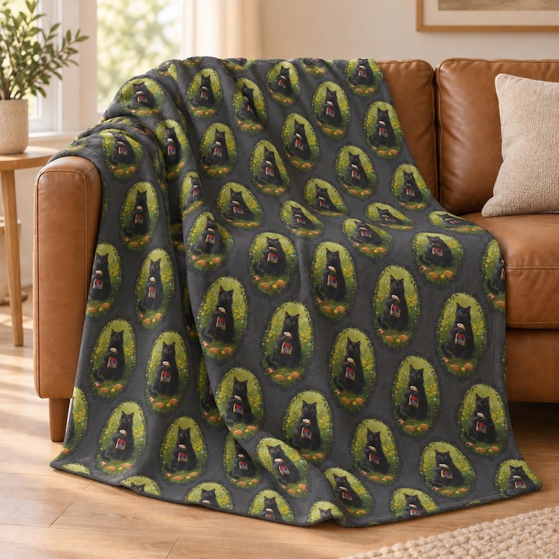 May include: A dark gray fleece throw blanket featuring a repeating pattern of black cats sitting inside oval frames. Each cat is depicted holding a small red mug, surrounded by a border of yellow flowers and green leaves against a dark background.