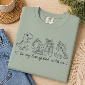 May include: Sage green Comfort Colors t-shirt with a graphic of a dinosaur, excavator, ballet shoes, and unicorn. The text "in my best of both worlds era" is also printed on the shirt.