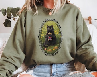 May include: Olive green sweatshirt featuring a framed illustration of a black cat holding a fast food bag. The cat is surrounded by yellow flowers and mushrooms. The sweatshirt is made of a soft material and has a crew neck.
