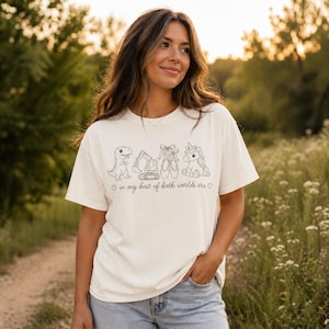 May include: Cream-colored t-shirt featuring a line drawing of a dinosaur, excavator, ballet shoes, and unicorn. The text "in my best of both worlds era" is below the drawings. The shirt is worn by a person in a field.