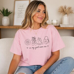May include: A light pink t-shirt with a black line drawing of a daisy, ballet shoes, a unicorn, and a heart-shaped purse. The text "in my girl mom era" is printed below the drawings.