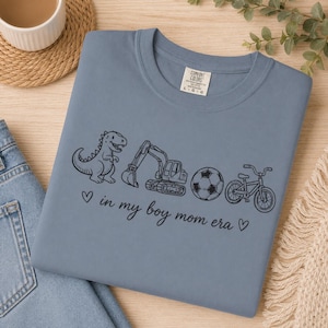 May include: A blue Comfort Colors t-shirt with a black graphic of a dinosaur, excavator, soccer ball, and bicycle. The text "in my boy mom era" is printed below the graphics.
