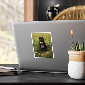 May include: A rectangular sticker featuring a black cat sitting in a field of yellow dandelions and green grass. The cat is holding a Taco Bell paper bag with both front paws. Two additional Taco Bell food items are visible on the grass nearby.