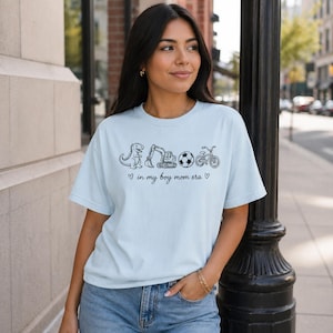 May include: Light blue t-shirt with a black graphic of a dinosaur, excavator, soccer ball, and bicycle. The text "in my boy mom era" is below the graphics. The shirt is made of a soft material.