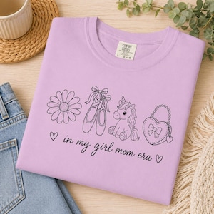 May include: A lilac-colored t-shirt with a black graphic design featuring a daisy, ballet shoes, a unicorn, and a heart-shaped purse. The text "in my girl mom era" is also printed on the shirt.