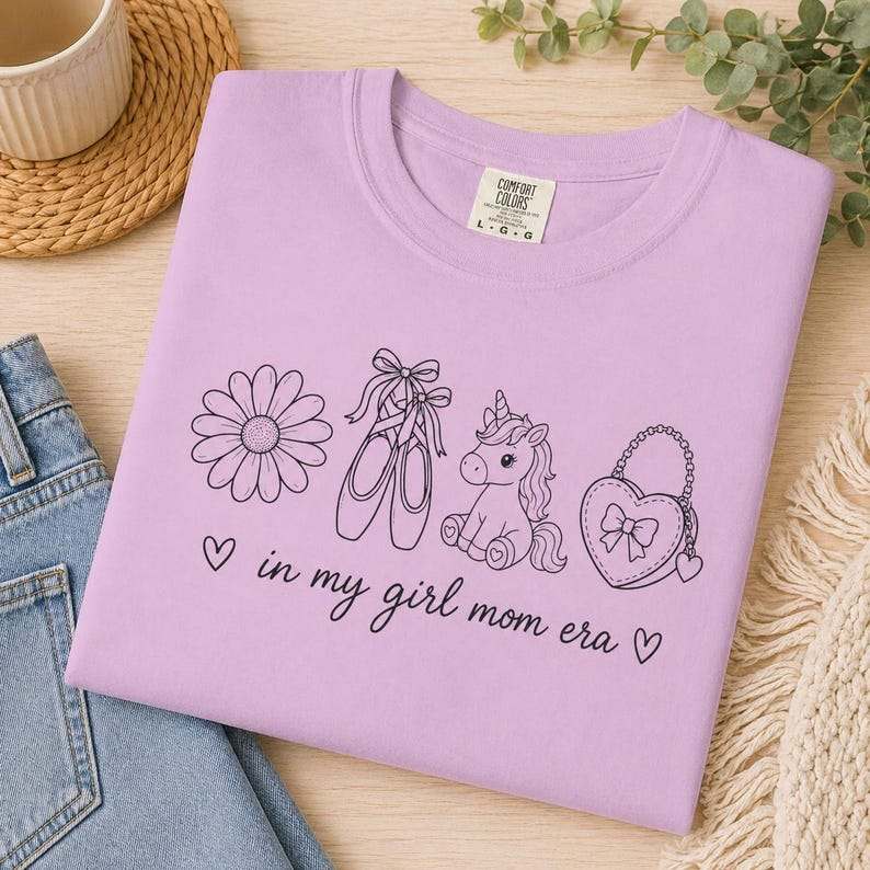 May include: A lilac-colored t-shirt with a black graphic design featuring a daisy, ballet shoes, a unicorn, and a heart-shaped purse. The text "in my girl mom era" is also printed on the shirt.