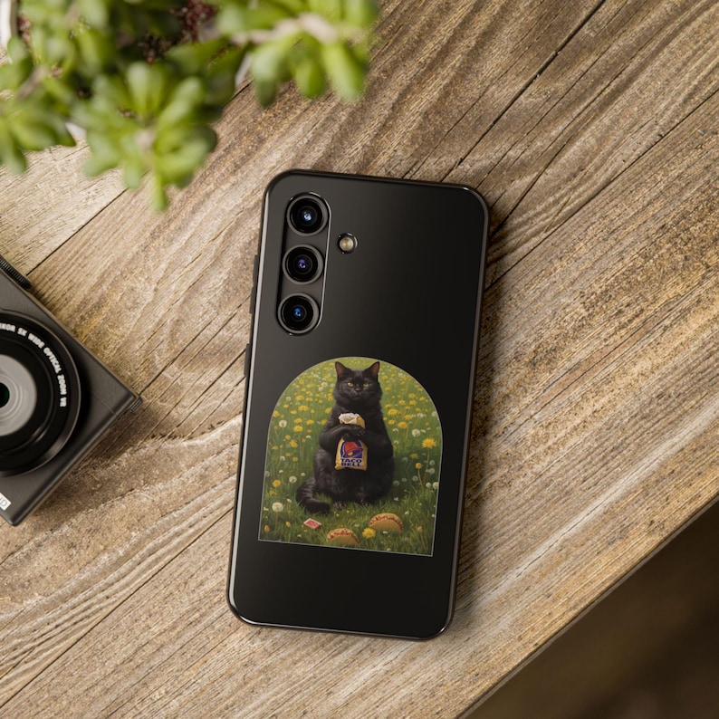 May include: A black smartphone case featuring a graphic of a black cat sitting in a field of yellow flowers, holding a Taco Bell bag with two tacos on the ground. The case has a vertical camera array with three lenses and a flash on the upper left side.