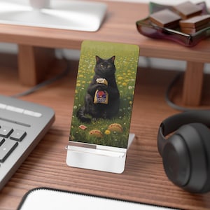 May include: A rectangular phone stand featuring a printed image of a black cat sitting in a field of yellow flowers and green grass. The cat is holding a Taco Bell paper bag, and two tacos are visible on the ground near the cat's feet.