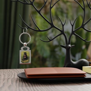 May include: A rectangular silver-toned metal keychain featuring a printed graphic of a black cat sitting in a field of yellow flowers. The keychain includes a circular split ring attached to the top, allowing it to hang from a metal hook.