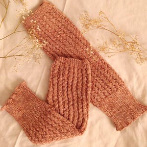 Puede incluir: Un par de calentadores de piernas de punto y un par de calcetines a juego en un suave color melocotón. Los calentadores de piernas tienen un patrón texturizado, mientras que los calcetines presentan un diseño acanalado. Los artículos están dispuestos sobre una superficie de color crema.