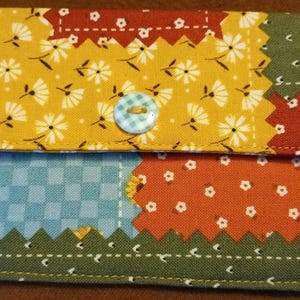 May include: A patchwork wallet with a button closure. The wallet features a variety of patterned fabrics, including yellow floral, blue gingham, and orange with white flower accents. A small loop is attached for carrying.