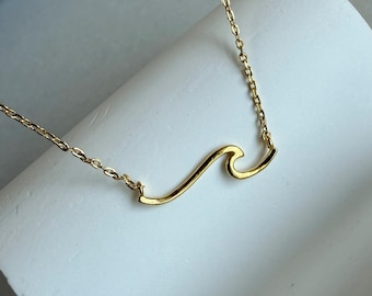 May include: A gold-colored necklace featuring a wave design. The pendant is a stylized wave shape, suspended from a delicate chain. The necklace is displayed on a white surface, highlighting its minimalist design.