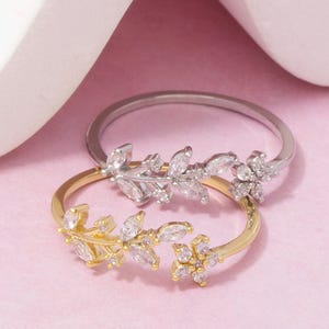 Könnte beinhalten: Zwei offene Ringe, einer aus silberfarbenem und einer aus goldfarbenem Metall, jeweils mit einem zarten Zweigdesign, das mit klaren, im Marquise- und Rundschliff facettierten Zirkonia-Steinen in Blatt- und Blütenmustern verziert ist.