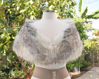 May include: A faux fur stole featuring a blend of white, gray, and brown tones with a thick, textured pile. The stole is draped over a white mannequin torso, showcasing a wide, V-shaped neckline and a soft, voluminous appearance throughout.
