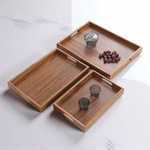 Könnte beinhalten: Drei rechteckige Serviertabletts aus Holz in verschiedenen Größen. Die Tabletts haben eine natürliche Holzmaserung und integrierte Griffe. Zwei kleine Gläser und eine kleine Glasschale befinden sich auf den Tabletts, zusammen mit einem Haufen dunkler Trockenfrüchte.