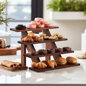 Puede incluir: Un soporte de madera oscura de tres niveles que exhibe una variedad de productos horneados. El nivel superior contiene donas glaseadas rosas y galletas de chocolate. El nivel medio tiene cruasanes y mini quiches. El nivel inferior presenta donas de chocolate, cruasanes y mini sándwiches.