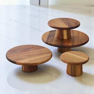 May include: A collection of three wooden cake stands in different sizes. The stands are crafted from light brown wood, showcasing a natural grain. Each stand features a circular top and a short, cylindrical base. They are displayed on a white surface.