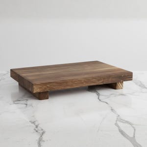 Puede incluir: Tabla de servir rectangular de madera marrón oscuro con base elevada. La madera presenta un veteado natural. La base consta de cuatro pequeños pies cuadrados. Diseñada para servir alimentos o exhibir objetos, mide unos 30 cm de largo.