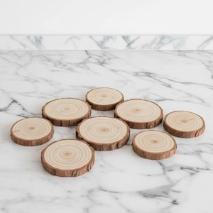 Könnte beinhalten: Eine Sammlung von acht Untersetzern aus natürlichen Holzscheiben. Jeder Untersetzer ist rund mit einer hellbraunen Mitte und einem dunkleren braunen Rindenrand. Die Untersetzer sind auf einer weiß-grauen Marmoroberfläche angeordnet.