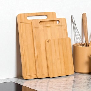 May include: Three bamboo cutting boards of varying sizes, leaning against a white wall. The boards have a natural wood grain finish and rectangular shapes. Two boards have handle cutouts, and one has a small hole. Kitchen utensils are in the background.