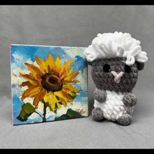 Peut inclure: Une petite peinture carrée d'un tournesol jaune sur fond de ciel bleu avec des nuages blancs, à côté d'un mouton crocheté gris et blanc. Le mouton a une tête blanche duveteuse et un corps gris.