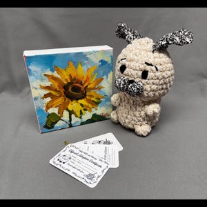 Peut inclure: Une petite peinture sur toile d'un tournesol aux pétales jaunes et au centre brun, sur fond de ciel bleu. À côté, un carlin crocheté beige avec des accents noirs et blancs. Des étiquettes de certificat d'adoption officielles sont au premier plan.