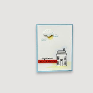 Peut inclure: Une carte de vœux rectangulaire avec une bordure bleu clair présentant un dessin au trait noir d'une maison, un soleil jaune et des nuages bleus. Un nuage blanc en relief est placé au-dessus du soleil. Le texte indique congratulations ON YOUR NEW HOME.
