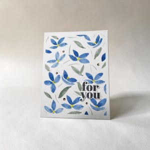 Peut inclure: Une carte de vœux blanche avec un motif aquarelle de fleurs bleues et de feuilles vertes. Le texte "for you" est imprimé dans une police sans empattement sombre et grasse dans le coin inférieur droit. De petits accents dorés sont dispersés.