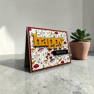 Peut inclure: Une carte d'anniversaire faite à la main avec le mot "happy" en lettres orange. La carte présente un fond à motifs avec des champignons et du feuillage. Une petite bannière noire indique "birthday". Une plante succulente dans un pot en terre cuite est à côté de la carte.