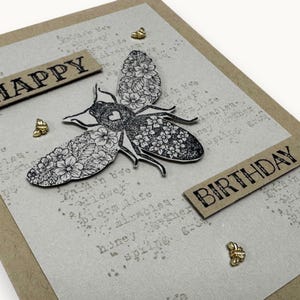 Peut inclure: Une carte d'anniversaire artisanale avec un motif d'abeille. Les ailes de l'abeille sont ornées de motifs floraux. La carte porte les mots "HAPPY" et "BIRTHDAY" en lettres noires. Des embellissements dorés en forme d'abeille sont dispersés.