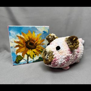 Peut inclure: Un petit cochon en peluche rose, blanc et marron est posé à côté d'une petite peinture d'un tournesol. Le cochon a des yeux noirs et un museau arrondi. La peinture représente un tournesol jaune vif sur fond de ciel bleu.