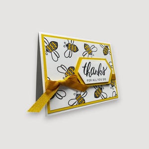 Peut inclure: Carte de vœux faite à la main avec une bordure jaune et un fond blanc orné d'abeilles noires et jaunes. La carte présente un ruban jaune et un hexagone blanc et jaune avec les mots "thanks for all you do."